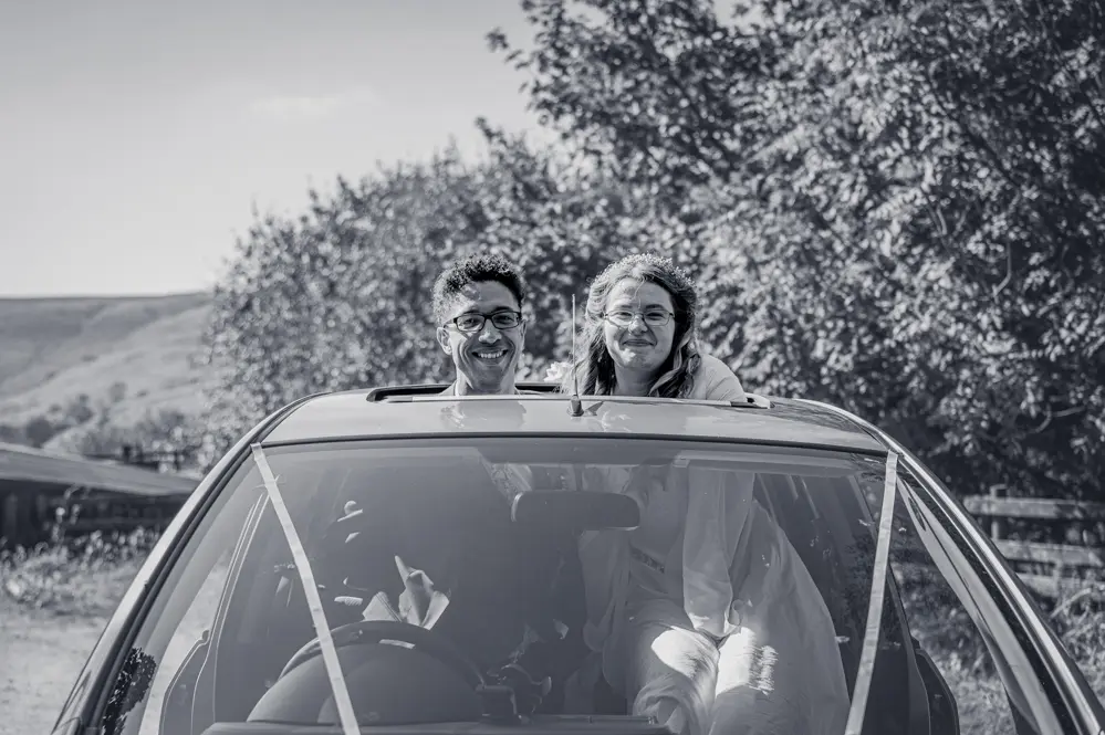 Bride and groom poking their heads through the sunroof of their wedding car on arrival at Cronkshaw Fold Farm, Rossendale