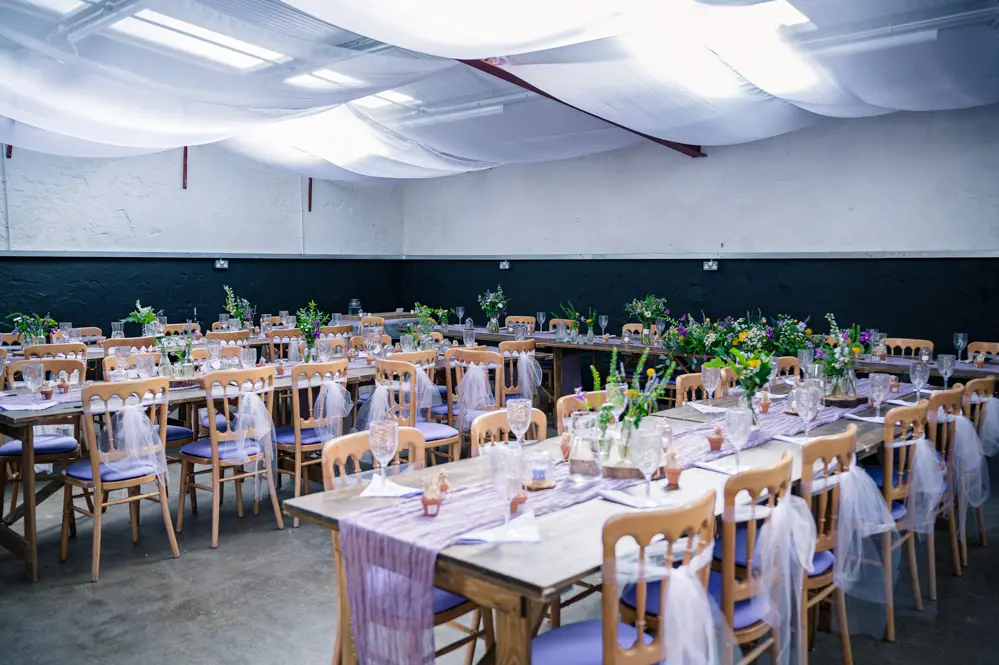 Overview of the Wedding reception decorations with purple cloth down the centre of tables and centre pieces with vases with wild flowers in them.