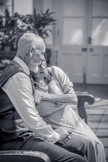 Black-and-white image of Lady resting head on man's shoulder and holding hands while sitting on a bench in a botanical gardens