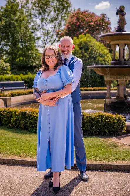 Man wrapping his arms round the front of his fiance with water fountain behind them