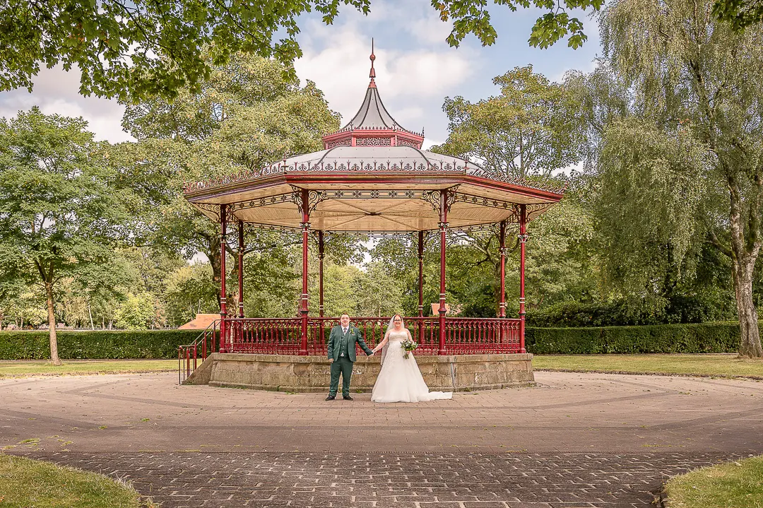 Bride and groom holding hands with the bandstand at Broadfield Park in Rochdale behind them