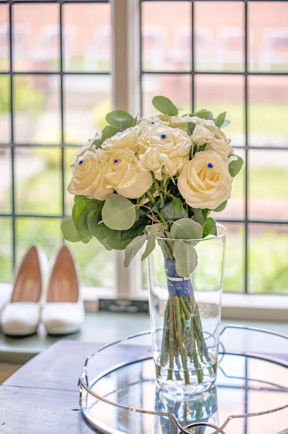 Bouquet in a vase on a mirrored tray with bridal shoes in the background