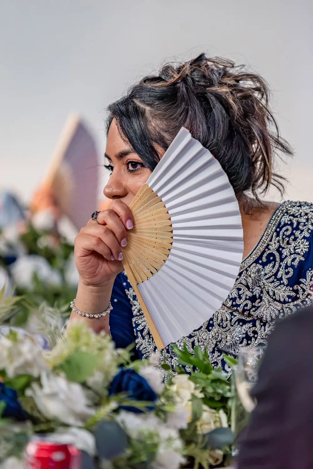 Candid shot of a bridesmaid in a blue dress using a hand fan to stay cool