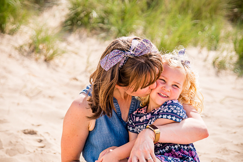 Outdoor Family Portrait of mother hugging and kissing daughter on the cheek while on the beach