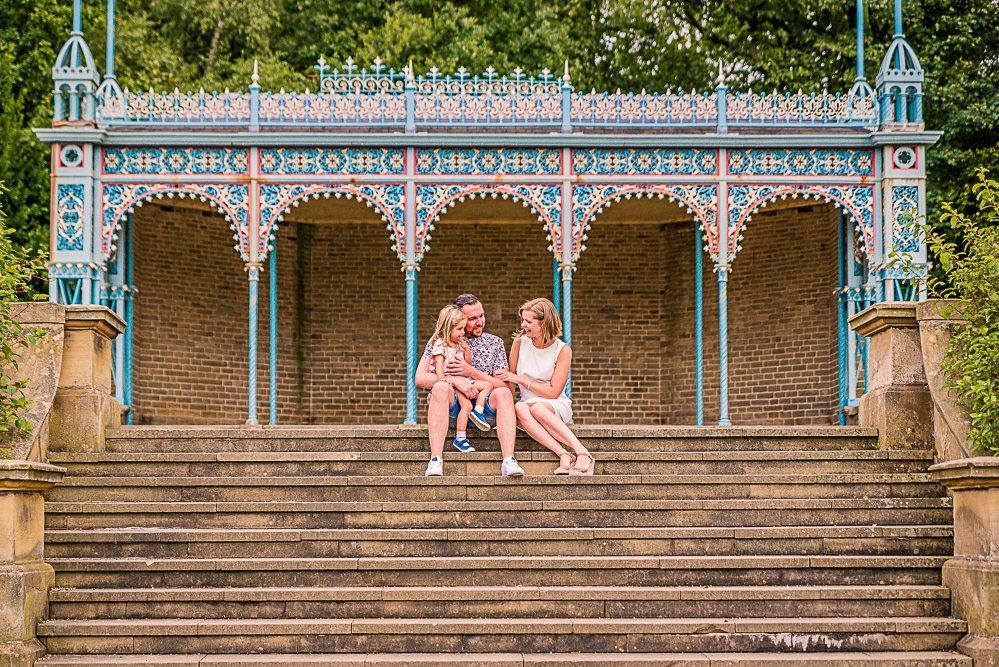 Outdoor Family Portrait of family of 3 seating on the top of stairs with a blue bandstand behind them