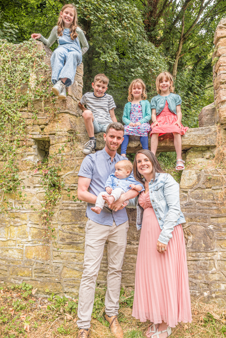 Outdoor Family Portrait of young family of 7 on while 4 of the children sit on a wall behind mum and dad hold baby.