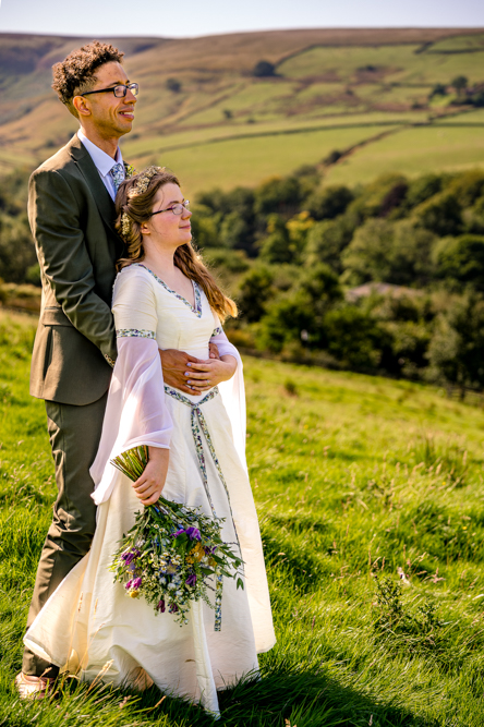 Bride and Groom holding hands in a field with hills behind. With Groom from behind