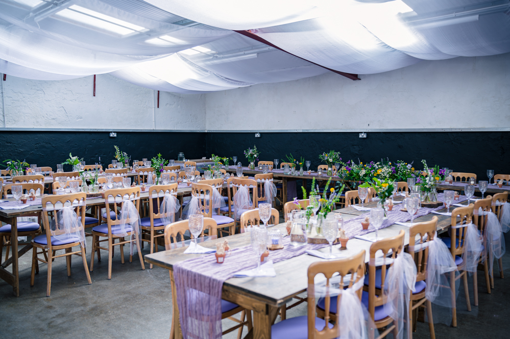 Overview of the Wedding reception decorations with purple cloth down the centre of tables and centre pieces with vases with wild flowers in them.