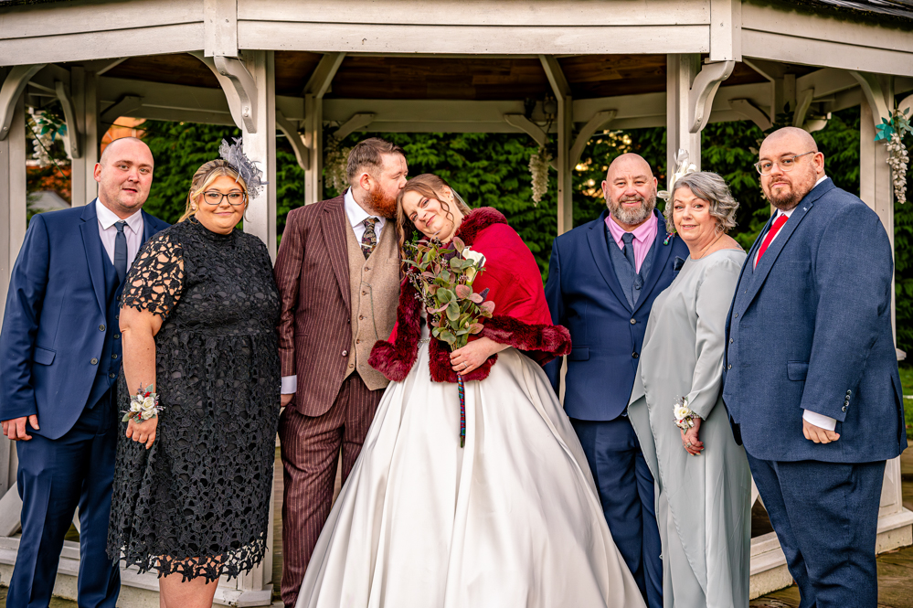 Groom kissing Brides head while picture being taken with grooms family with bandstand behind them