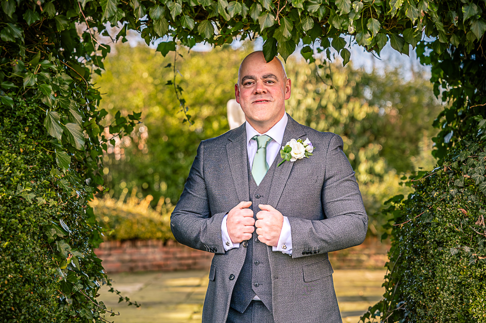 Groom under a leaf shaped arch holding his jacket with each hand