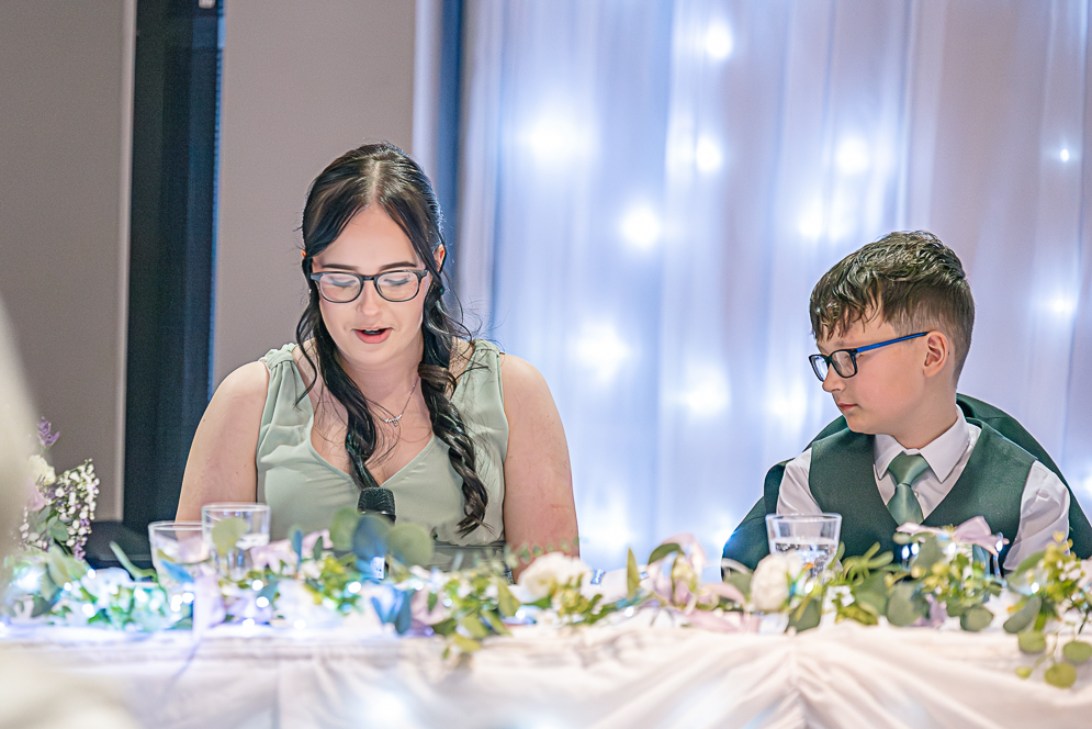 View of groomsman (boy) listing to bridesmaid (girl) giving a speech