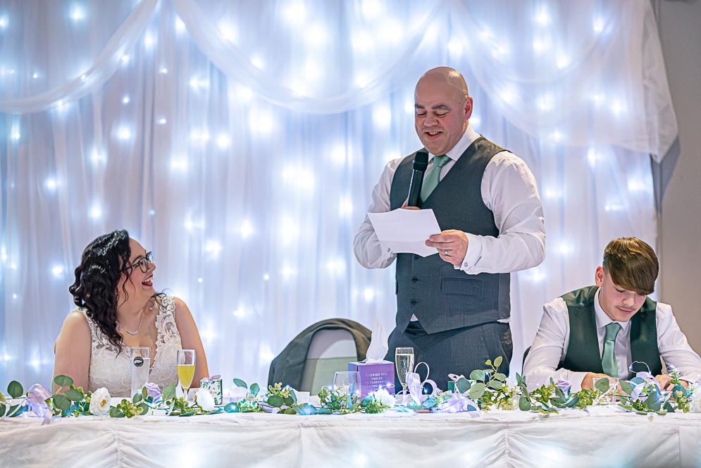 Groom’s speech, bride smiling and groomsman listening.