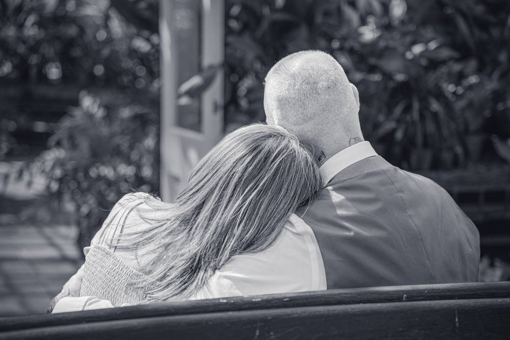 Black-and-white image of The view from behind as Lady resting head on man's shoulder and holding hands while sitting on a bench in a botanical gardens