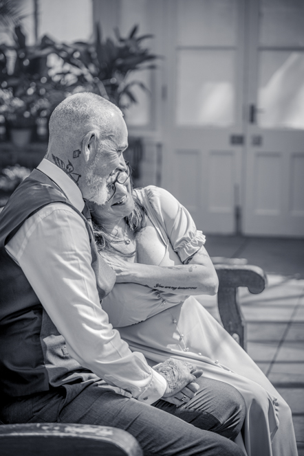 Black-and-white image of Lady resting head on man's shoulder and holding hands while sitting on a bench in a botanical gardens