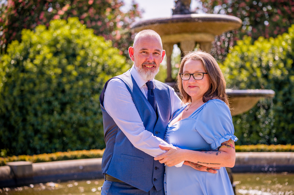 Close up a couple with arms around each other with water fountain behind them