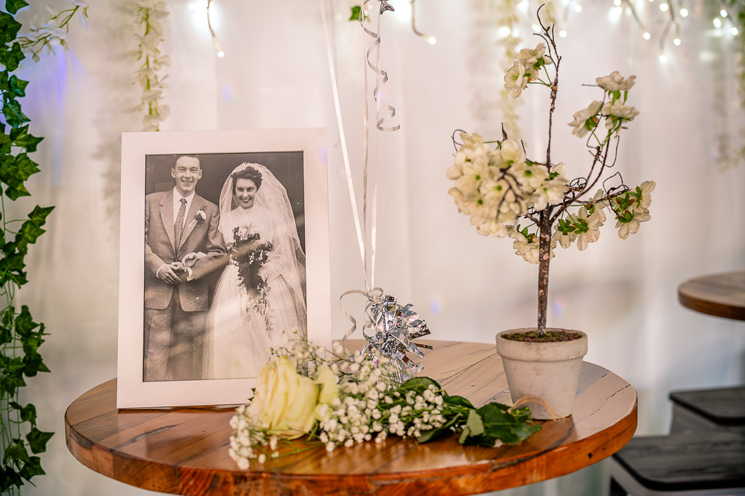 Framed black and white wedding photograph displayed on a reception table with bouquet and white potted plant.