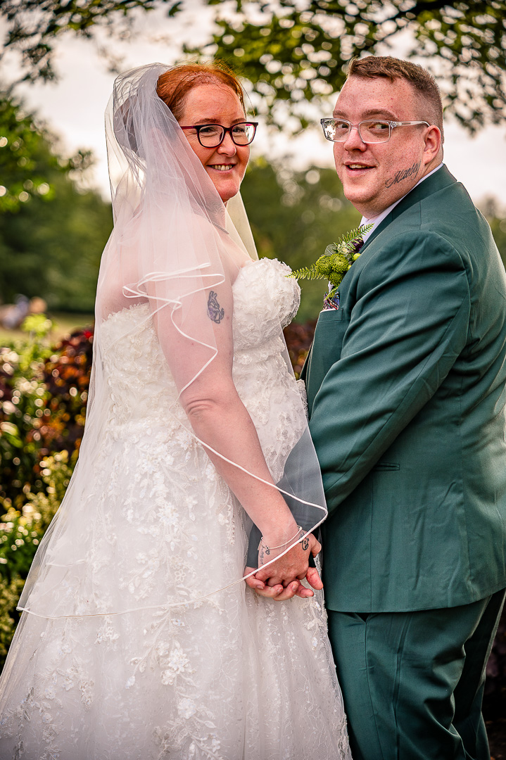 Bride and groom standing hand in hand beneath the trees at Broadfield Park in Rochdale
