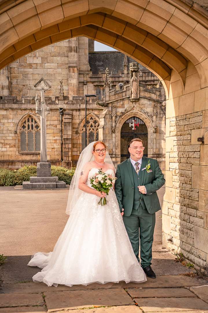 Bride and groom standing beneath a stone arch with St Chad’s Church visible behind them