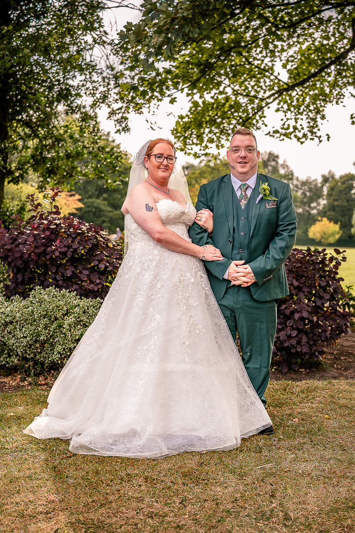 Bride and groom standing with brides hands armed grooms arm beneath the trees at Broadfield Park in Greater Manchester