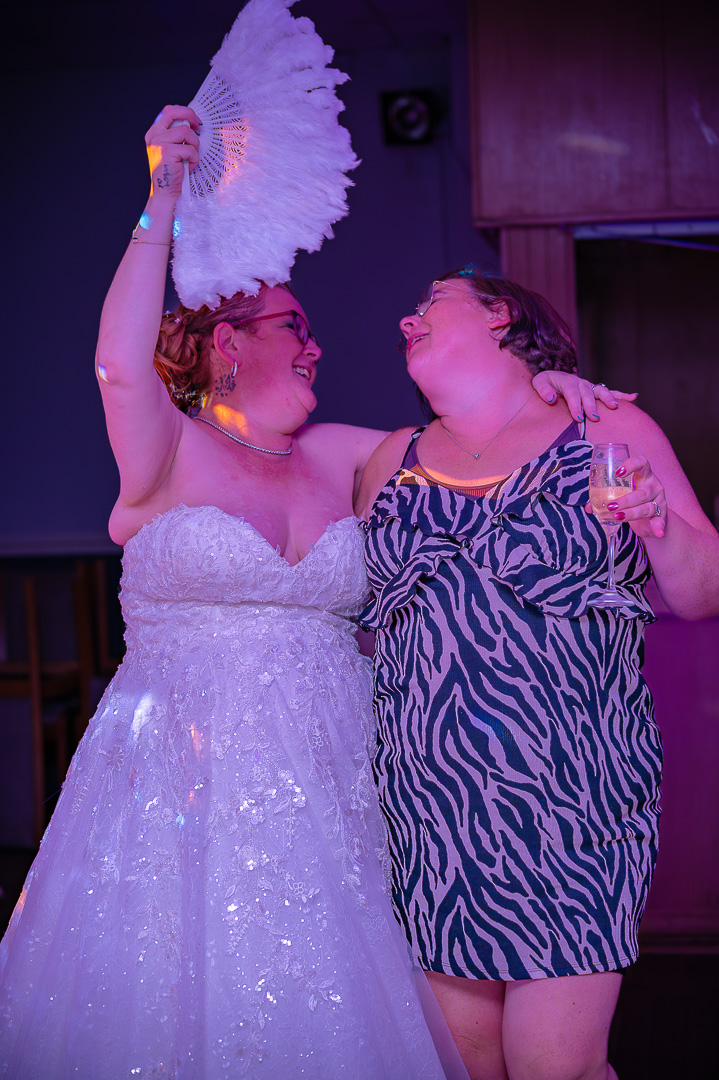 Bride holding a fan over her head while a guest looks on holding a glass, at an elegant and joyful wedding reception