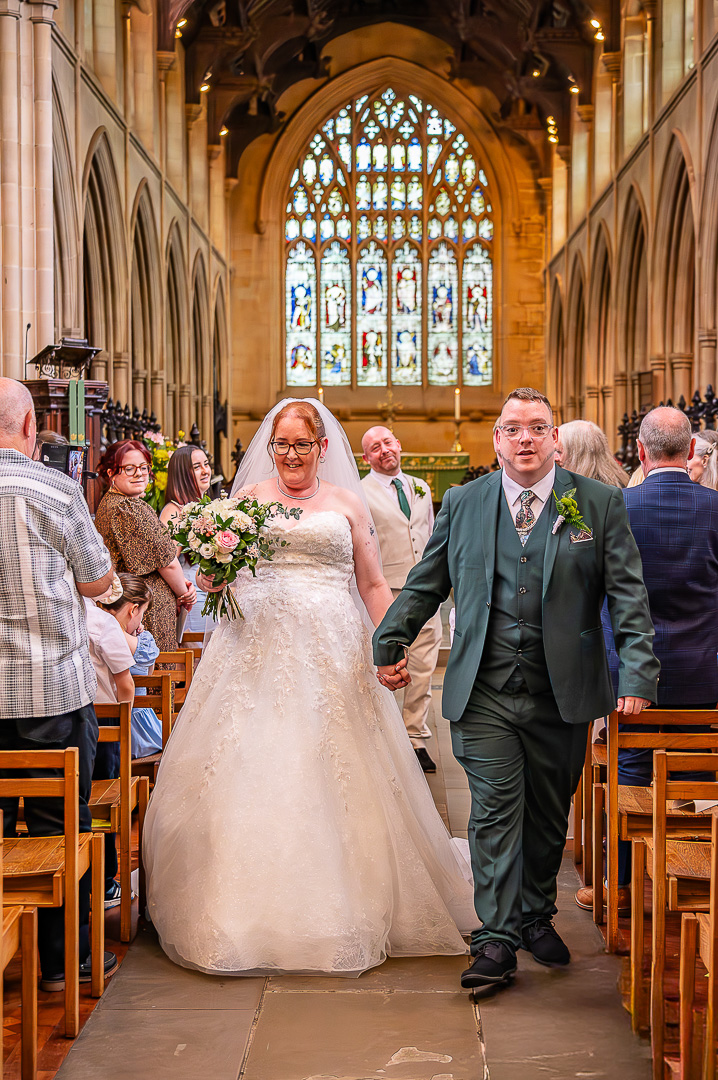Bride and groom walking back down the aisle with the bride’s brother smiling in the background at St Chad’s Church, Rochdale during their elegant Christian wedding