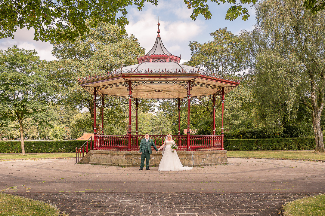 Bride and groom holding hands with the bandstand at Broadfield Park in Rochdale behind them