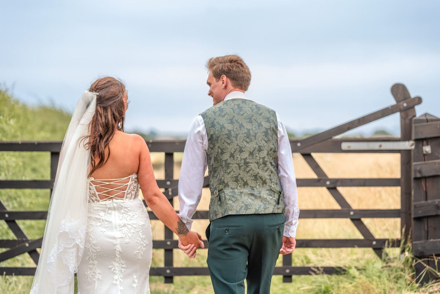 Bride and groom walking hand in hand towards a field gate at Denroyd Farm, Huddersfield, the groom glancing back at his bride